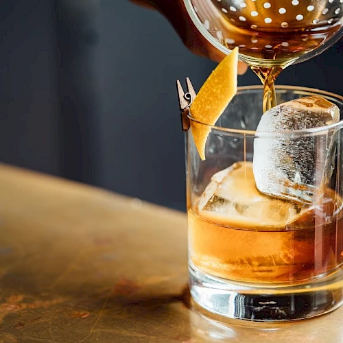 A glass of whiskey on the rocks with a large ice cube, a twist of lemon, and another drink being poured from a bottle.