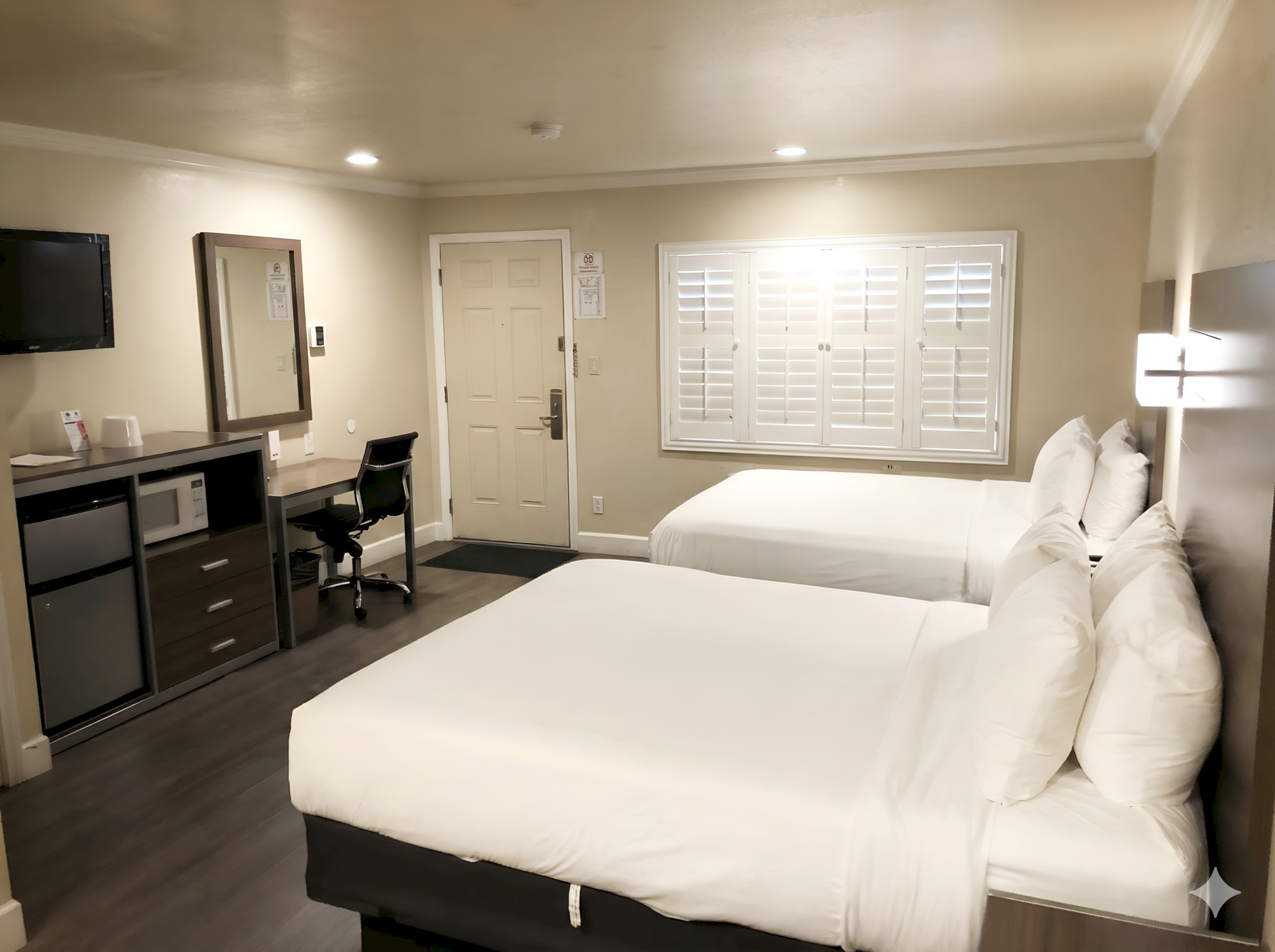 Two double beds in a hotel room with a desk, mini-fridge, flat-screen TV, and a window with shutters, all in a neat, modern setup.
