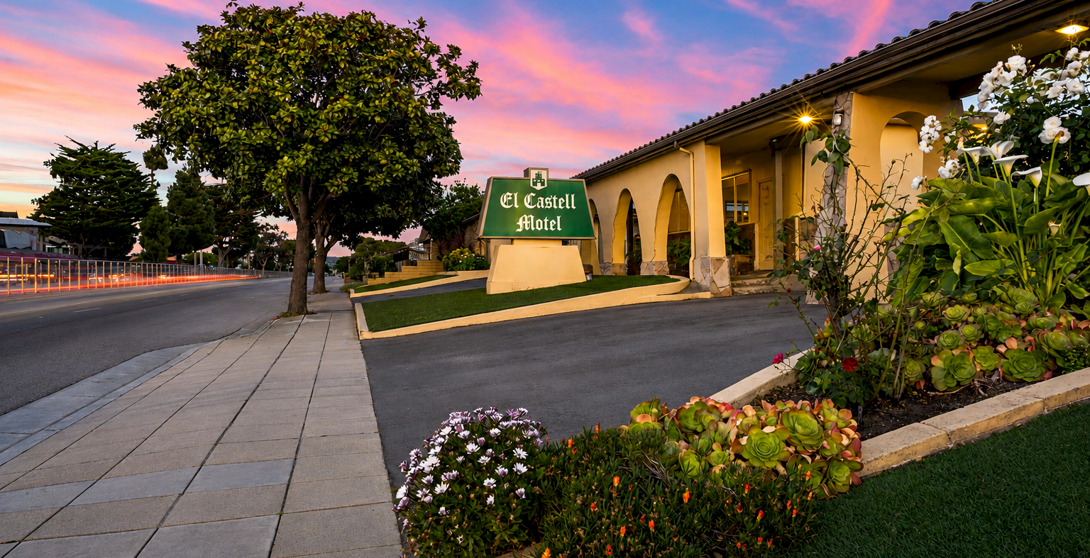 A calm hotel entrance at dusk with a paved walkway, flowering shrubs, and a green sign reading &ldquo;El Castillo&rdquo; under a pastel sky.