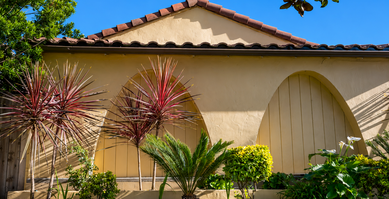 A beige stucco house with a tiled roof, arched doorway, and a row of spiky red-leaf plants plus palm and green shrubs in a neat front yard.