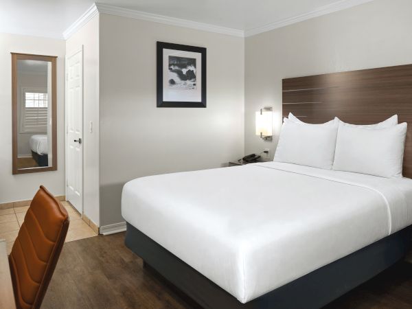 A clean hotel room with a king bed, white linens, wooden headboard, nightstands, a wall lamp, framed art, a small desk with a chair, and a doorway to a bathroom.