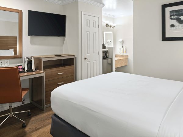 A bright hotel room with a white bed, wall TV, desk with coffee maker, armchair, artwork, and an open bathroom doorway.