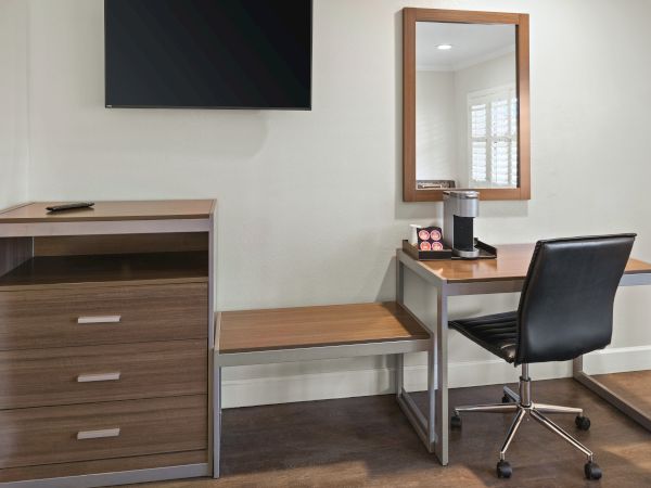 A modern hotel-style desk area with a wall-mounted TV, dresser, a desk with chair, mirror, and a coffee/tea station, all in a neat setup.