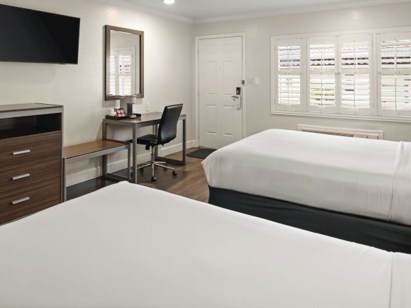 A hotel room with two queen beds, a desk and chair, a dresser with a mounted TV, a mirror, window blinds, and a doorway in a bright, modern space.