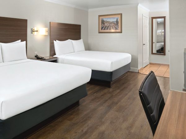Two hotel beds with white linens, dark wood headboards, a desk and chair, door to a hallway, and a wood floor in a tidy room.