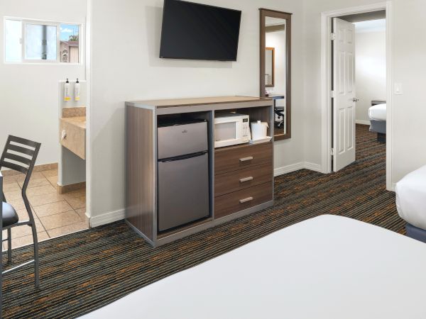 A hotel room with a TV mounted on the wall, a cabinet with a mini-fridge, drawers, and shelves, a doorway to a bathroom, and part of a bed in the foreground.