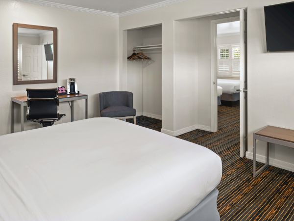 A hotel room with a bed in the foreground, a desk and chair, a wall mirror, a small seating area, and an open closet; doorway to another room is visible.