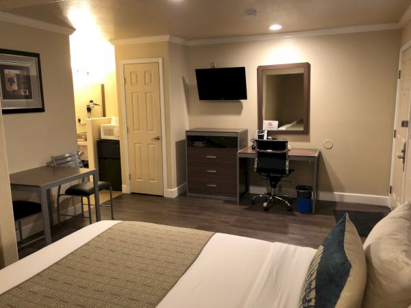 Hotel room with a bed, desk area, wall-mounted TV, dresser, mirror, and a small seating nook&mdash;neutral tones and modern furnishings.