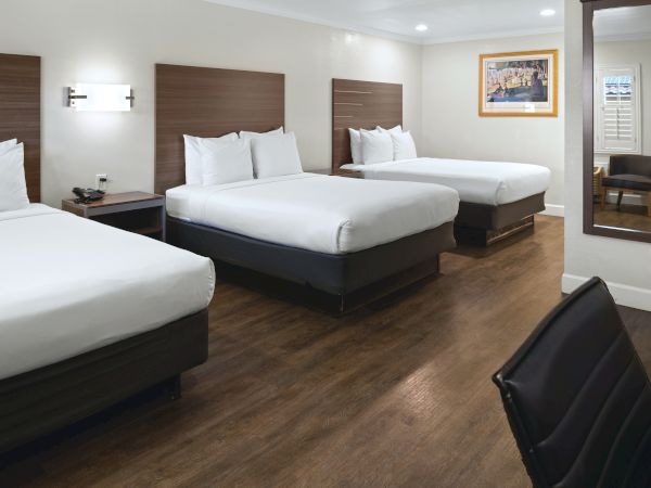 A hotel room with three queen beds, wooden headboards, white linens, bedside tables, a chair, and a full-length mirror in a clean, modern setup.
