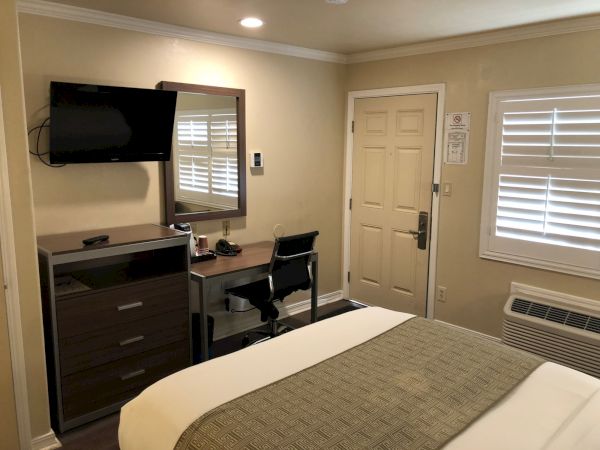A hotel room with a bed, dresser, wall-mounted TV, desk and chair, mirror, door, blinds, and an AC unit&mdash;neat and simple, ready for guests.