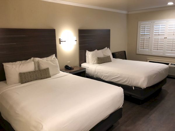 Two neatly made hotel beds with white linens, dark wood headboards, bedside lamps, and a window with shutters in a tidy room.