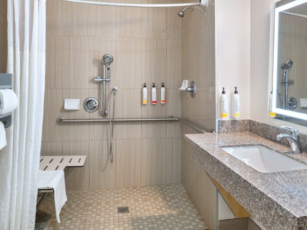 A wheelchair-accessible bathroom with a walk-in shower, grab bars, a bench, and a granite sink counter. The image shows a bathroom setup designed for accessibility.