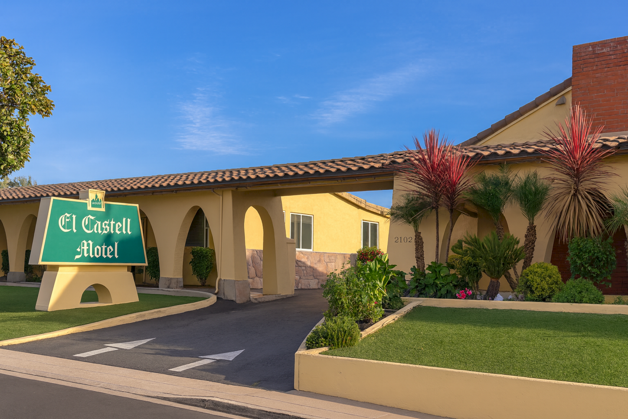 A single-story hotel with a green sign reading &ldquo;El Castell Motel,&rdquo; arched entrance, desert landscaping, and a sunny blue sky, end.