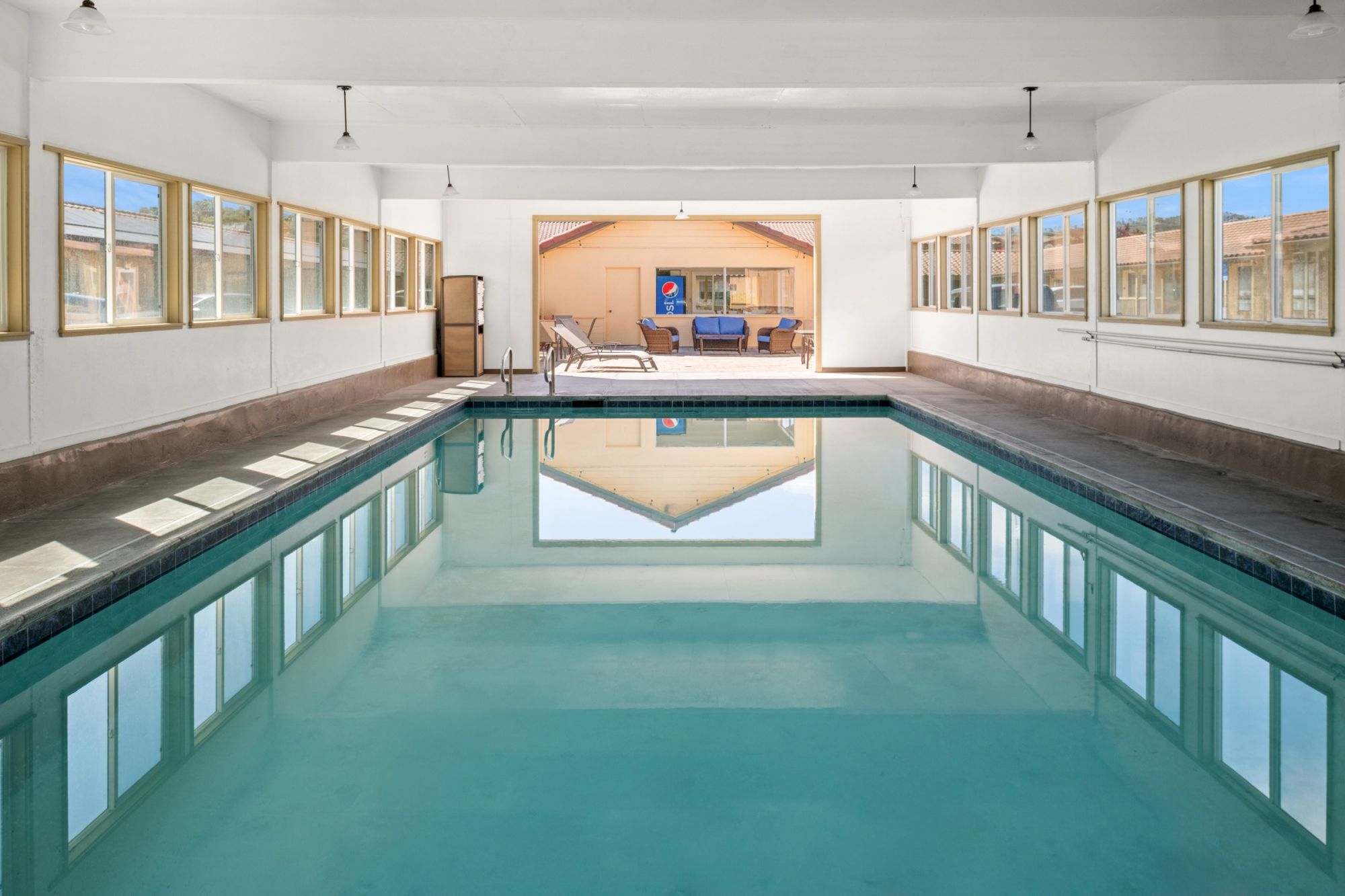 Indoor backyard with a long rectangular swimming pool, glass walls/windows, and patio seating under hanging lights.