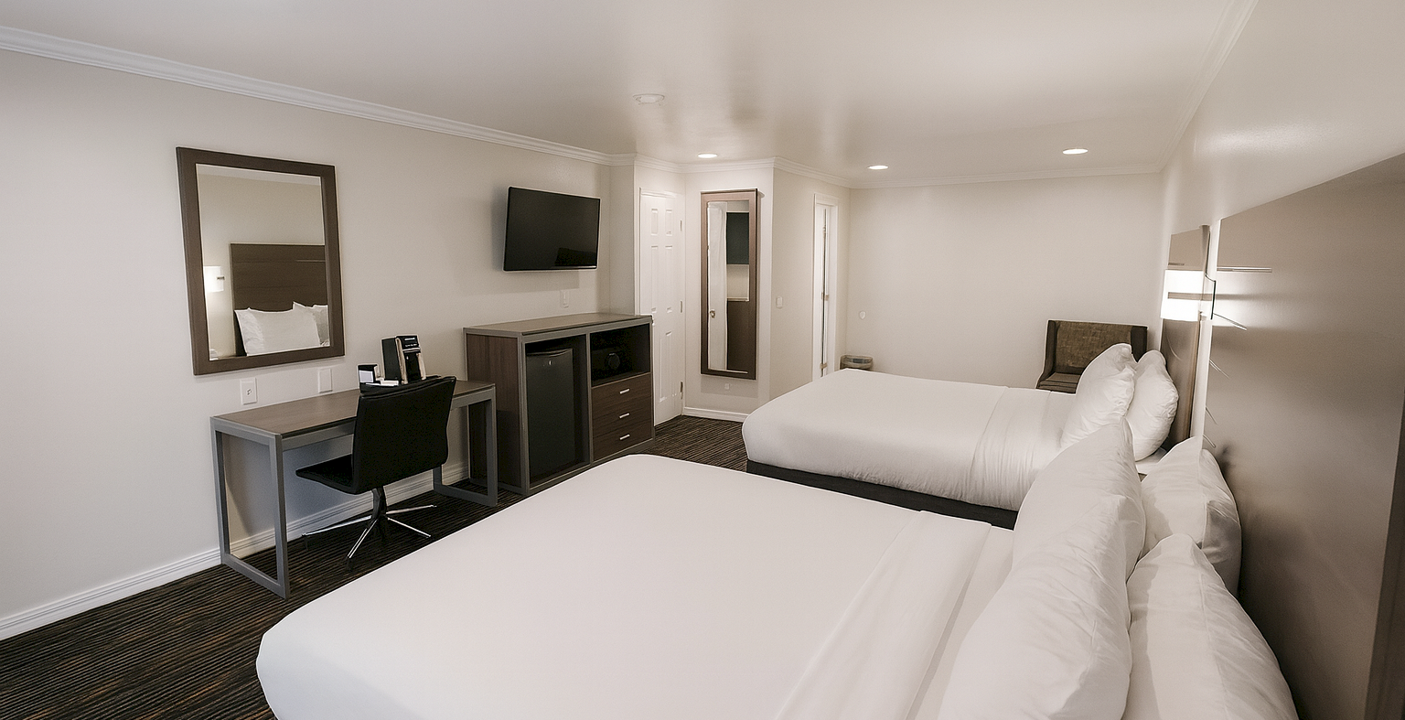 Two double beds in a clean hotel room with a work desk, chair, wall mirror, dresser with a TV, and a door leading to a private area.
