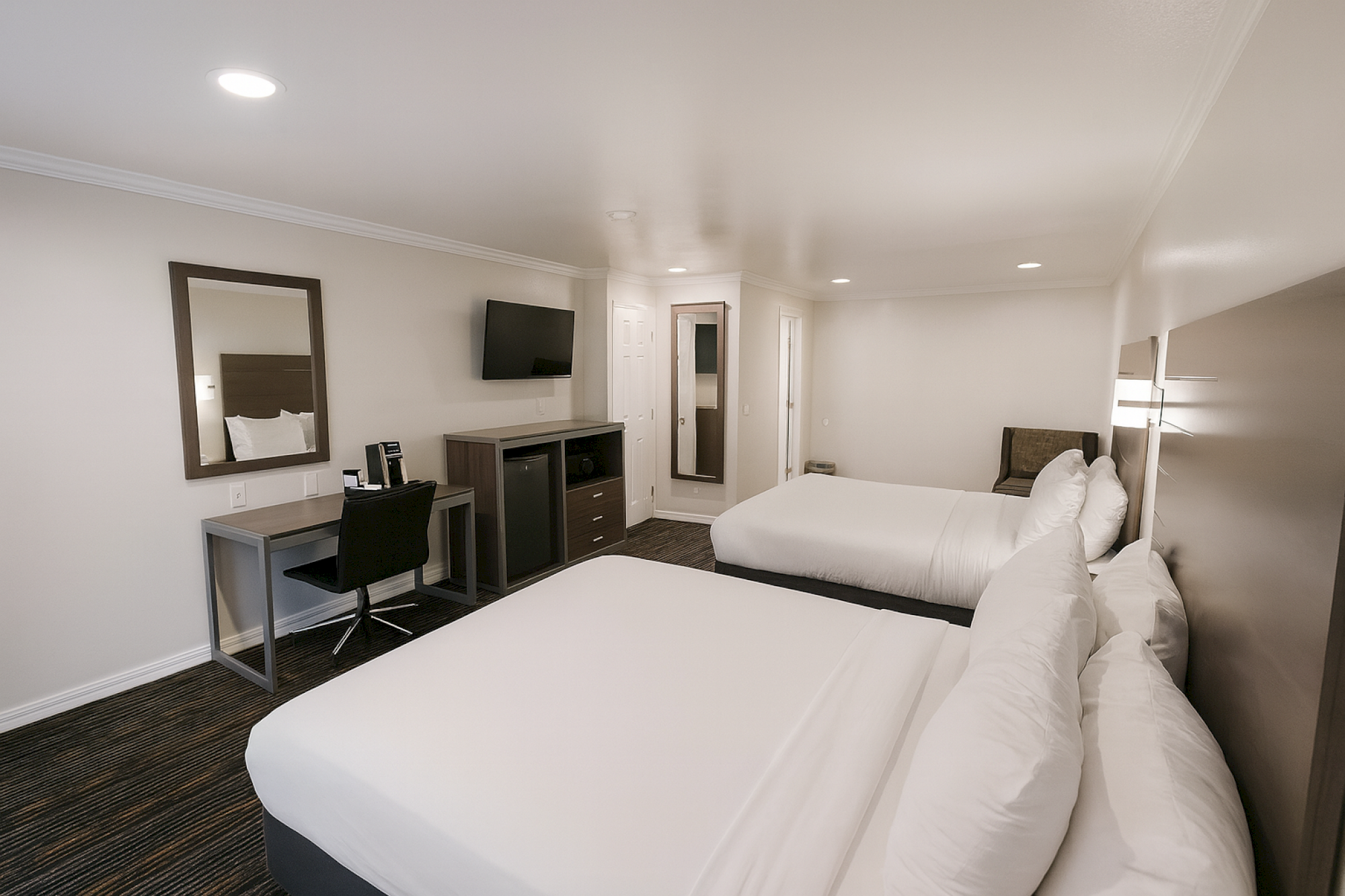 Two double beds in a clean hotel room with a work desk, chair, wall mirror, dresser with a TV, and a door leading to a private area.