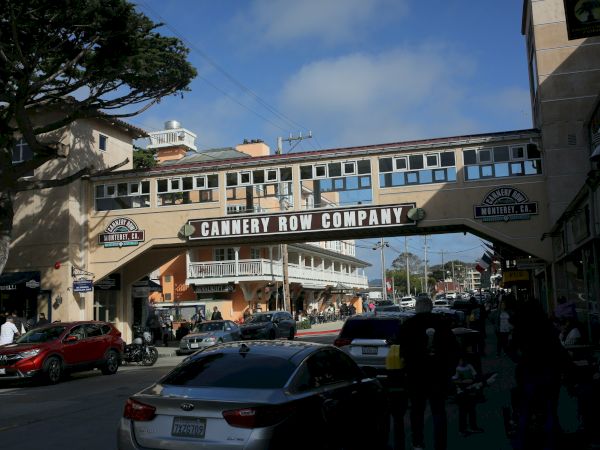 A sunlit street scene under a pedestrian bridge that reads &ldquo;CANERY ROW COMPANY,&rdquo; with cars, people, and buildings on either side.