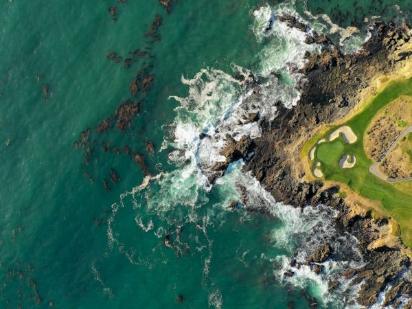 Aerial view of a rugged coastline: turquoise sea on the left, white waves breaking along dark rocks, and a grassy cliff with a winding shoreline on the right.