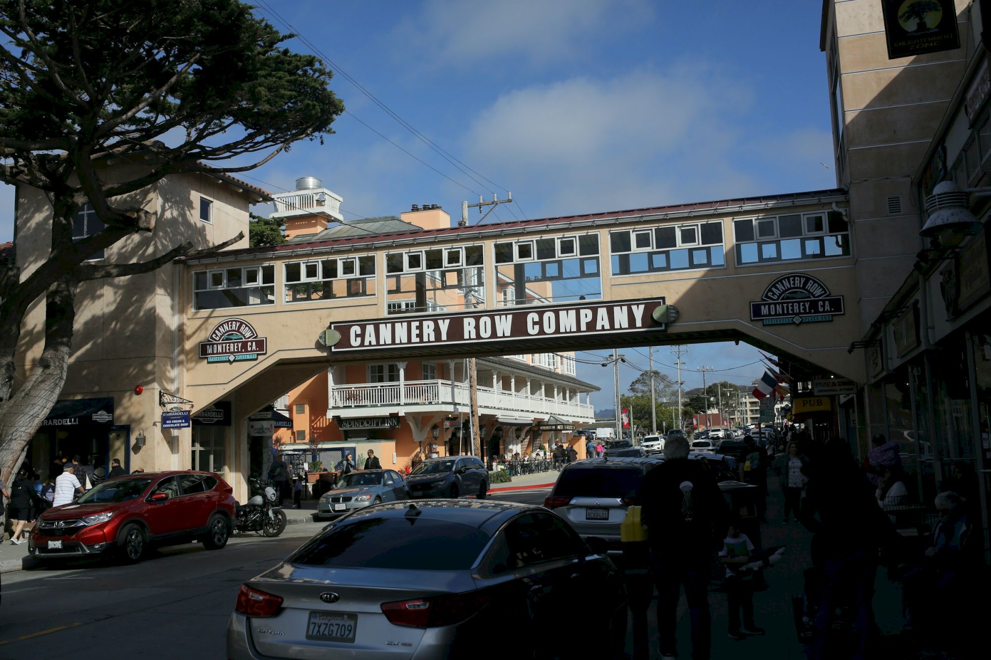 A busy street scene under a covered pedestrian bridge labeled &ldquo;Cannery Row Company,&rdquo; with cars, buildings, and pedestrians on a sunny day.
