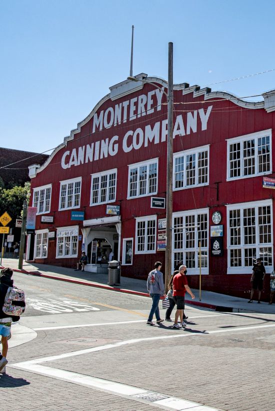 Colorful red brick building labeled &ldquo;Monterey Canning Company&rdquo; at a sunny street corner with pedestrians crossing.
