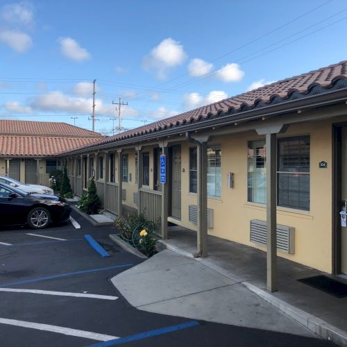 A row of motel rooms with blue awnings, a parking lot, and a bright blue sky overhead. B99.