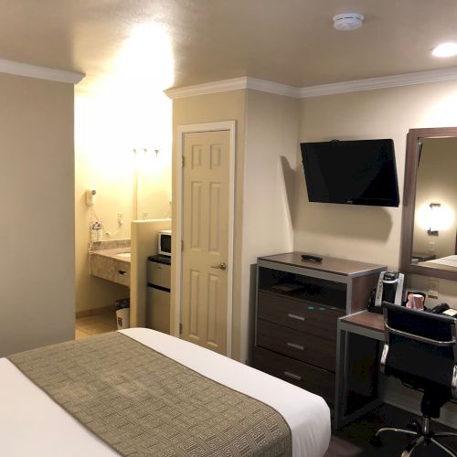 A small hotel room with a bed, desk, monitor, and flat-screen TV; neutral colors, wall art, and a door leading to a hallway.