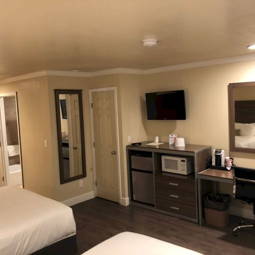 Two hotel rooms with beds, a desk, mini-fridge, microwave, wall-mounted TV, mirror, and a chair; neutral beige walls and modern decor.