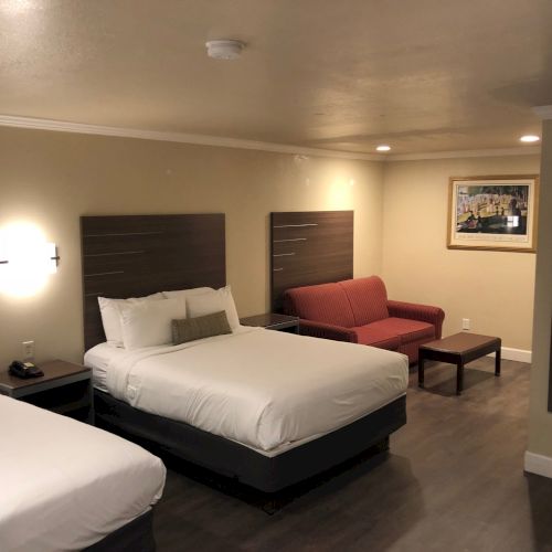 A hotel room with two queen beds, a red sofa, a coffee table, a nightstand, wall lamps, and a small workspace; warm lighting and neutral decor.