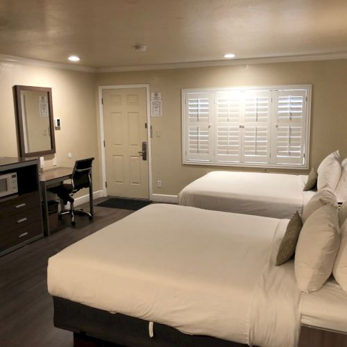 A hotel room with two queen beds, a desk and chair, a mini-fridge, a wall-mounted TV, and a window with shutters.