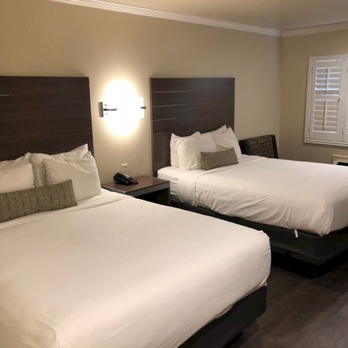 Two neatly made hotel beds with white linens and headboards, a shared nightstand, lamps, and a window with shutters.