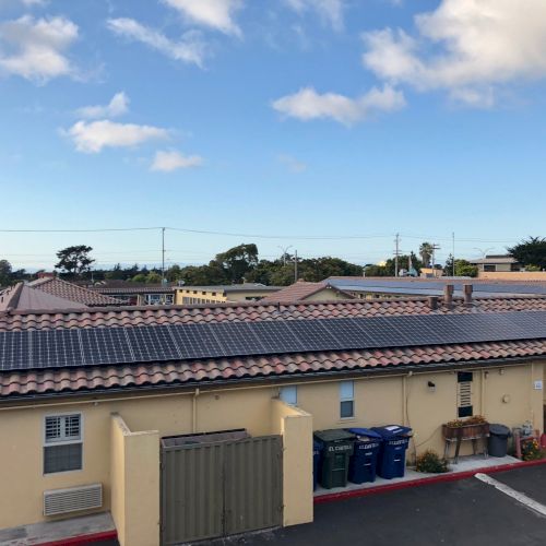 A small parking area next to a row of single-story buildings with a blue sky and clouds above.