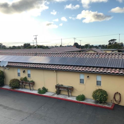 A sunny day scene at a small outdoor market or storefront, with a long low building, potted plants, a parking lot, and blue sky with clouds, end.