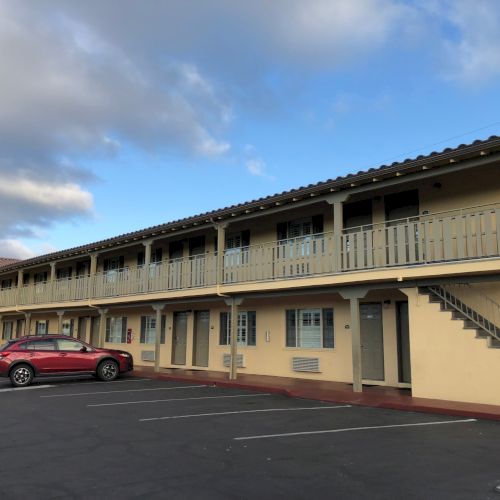 A two-story motel with yellow walls, brown trim, and a row of doors; a red car parked outside, sunny day, clear sky.