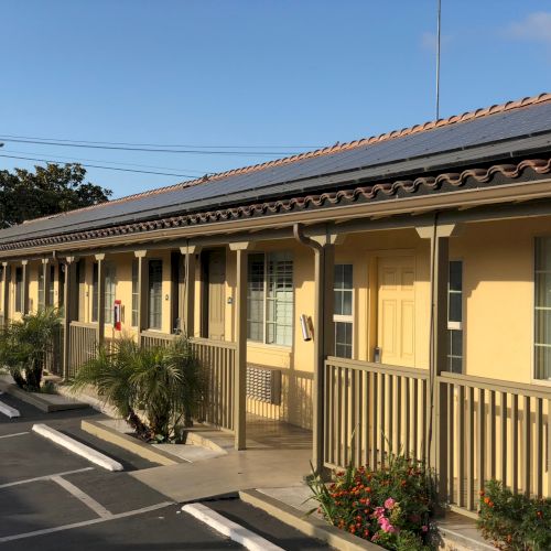 A single-story motel or hotel with a covered walkway, yellowish walls, and parked cars along the curb.