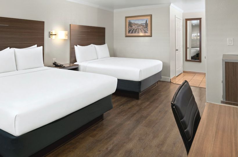 Two double beds in a modern hotel room with wood floors, white linens, a desk chair, a desk, and a doorway to a bath area.