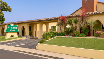 A single-story motel with a green sign reading &ldquo;El Capell Motel&rdquo; sits along a curved driveway, yellow walls, arches, palm trees, and well-kept landscaping.