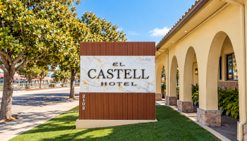 A sunny hotel entrance with a sign reading &ldquo;El Castelli Hotel&rdquo; beside landscaped grass, trees, and arched walkway.