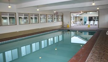 An indoor rectangular swimming pool with tiled edges, bright overhead lights, windows along one wall, and a lounge area in the background.