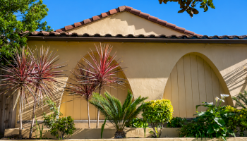 A beige stucco house with a red-tiled roof, arched door, and a small garden featuring red-tipped yucca, palm, and leafy shrubs; sunny day.