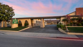 A low-rise hotel or motel with a covered drive-through entrance, a manicured front yard, palm trees, and a sunset sky in pink and blue hues.