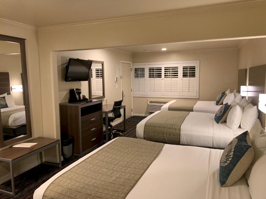 A hotel room with two queen beds, a desk, TV, dresser, and a small seating area, all in neutral tones with lamps and windows.