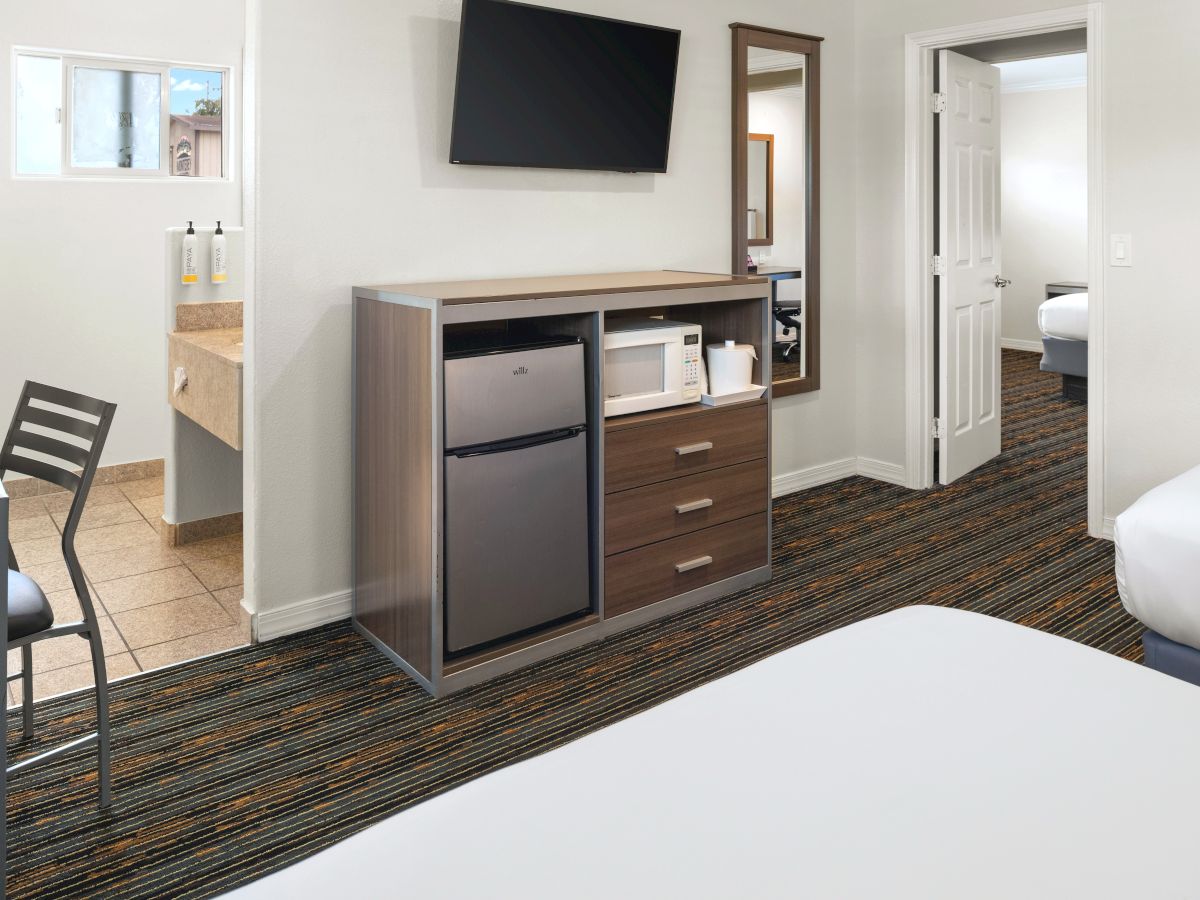 A hotel room with a TV on the wall, a cabinet housing a mini-fridge and drawers, a small desk, doorway to a bath, and a bed in the foreground.