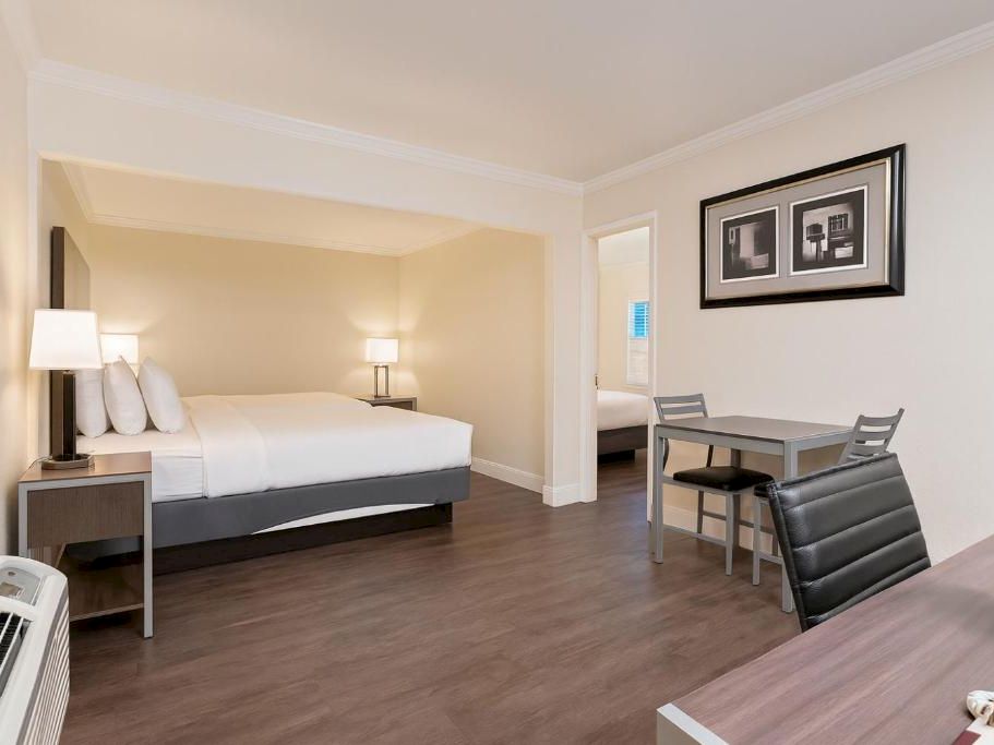 A hotel room with a king bed, two nightstands, a small work desk, a seating area, and a dining table; modern decor and wood floors.