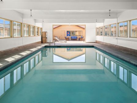An indoor swimming pool with turquoise water, white walls, long row of windows, and a seating area at the far end with a tv cabinet reflected in the calm water.