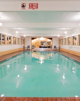 An indoor swimming pool with tiled deck, bright lights, and large windows along both sides, empty lounge area, calm water, reflective surfaces.