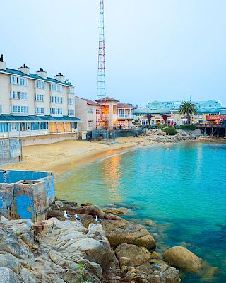 Colorful seaside village with pastel buildings, a rocky shore, clear turquoise water, and a pier extending into the calm sea.