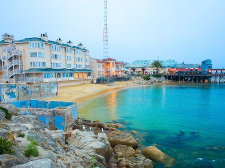Colorful seaside village with pastel buildings, a rocky shore, clear turquoise water, and a pier extending into the calm sea.