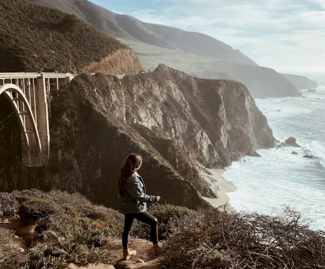 A person stands on a cliff edge near a railing bridge, overlooking rugged coastline and the sea under a cloudy sky.