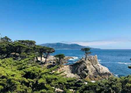 Cliffs, greenery, and rocks by the blue sea under a clear sky, a scenic coastal view with waves hitting the shore.