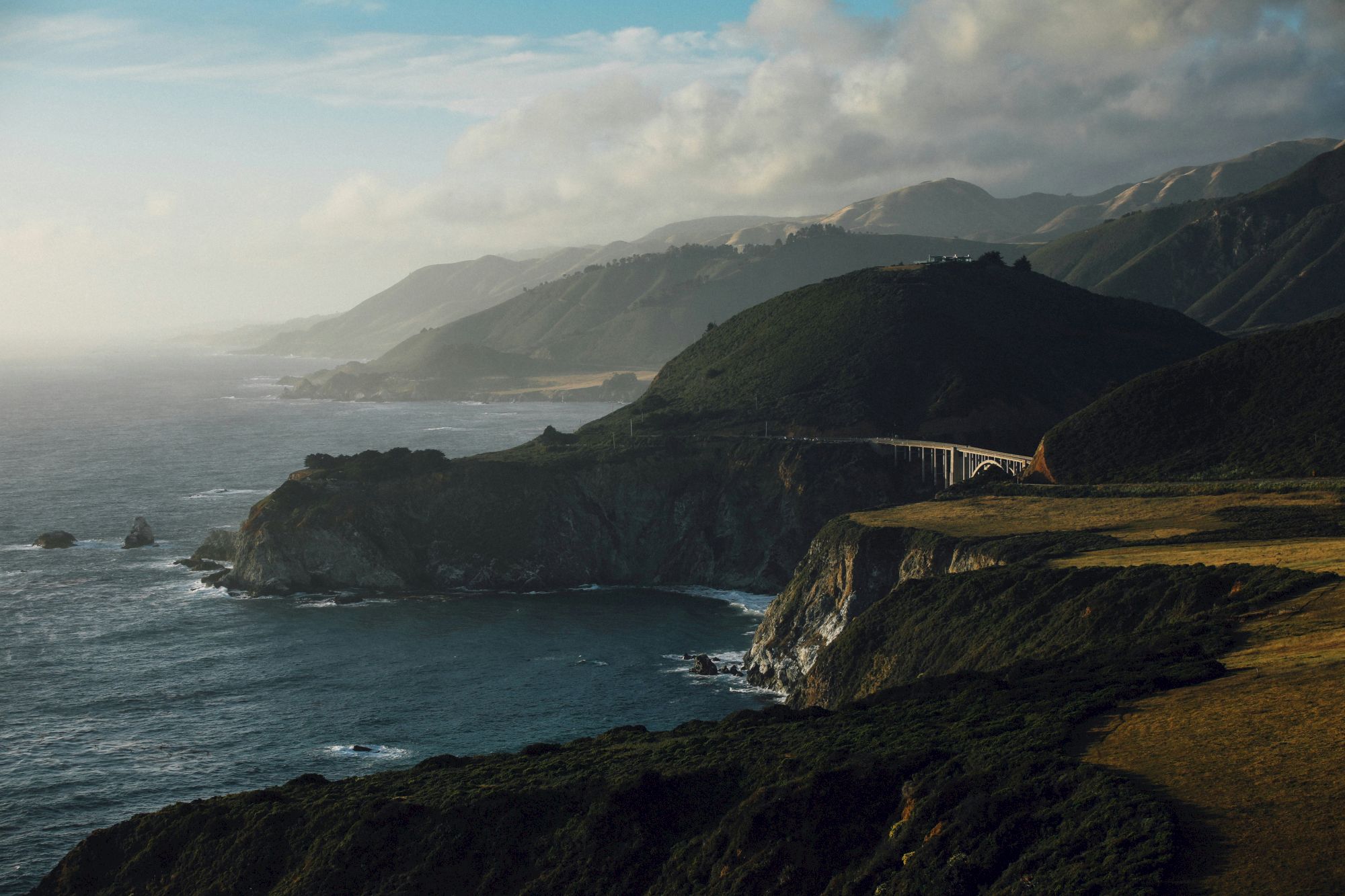 A rugged coastal scene with a cliffside road and arching bridge along green hills, waves crashing against rocks, and misty blue sea.